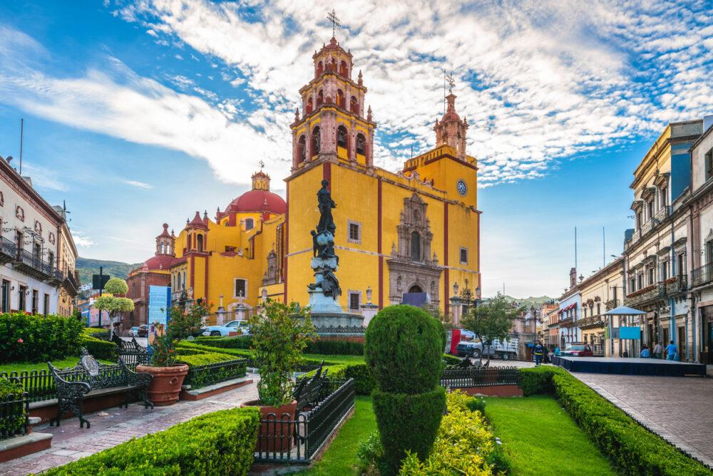Historic yellow cathedral with clock tower, surrounded by green gardens and benches, under a blue sky with clouds.