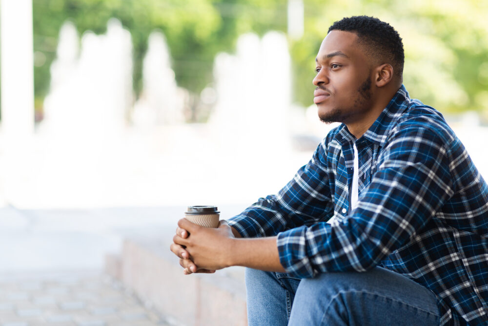 Man in plaid shirt sitting outdoors holding a disposable coffee cup, with blurred background trees and fountains.