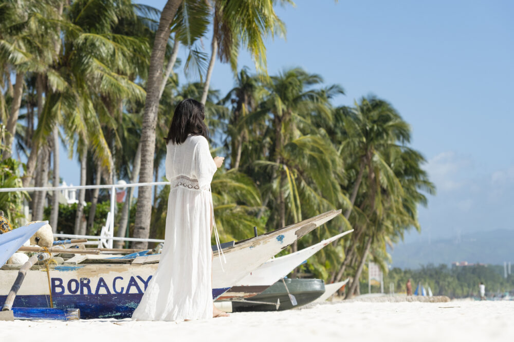 Woman in white dress stands near boats on tropical beach with palm trees.