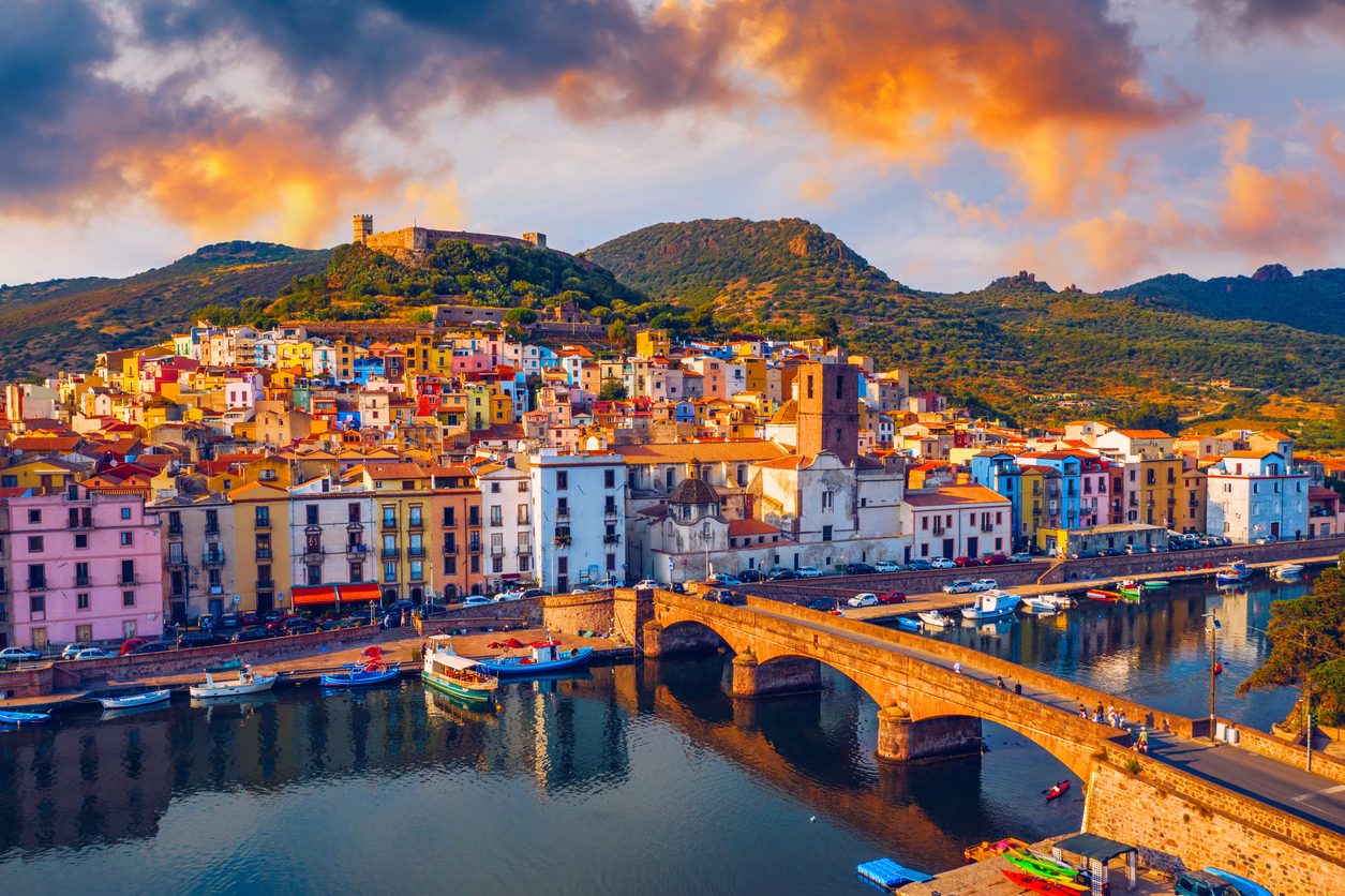 Colorful hillside town with stone bridge over water, boats docked, and mountains in the background at sunset.