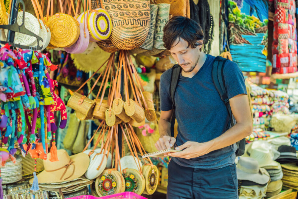 Man with backpack using tablet in colorful outdoor market with hats and bags displayed.