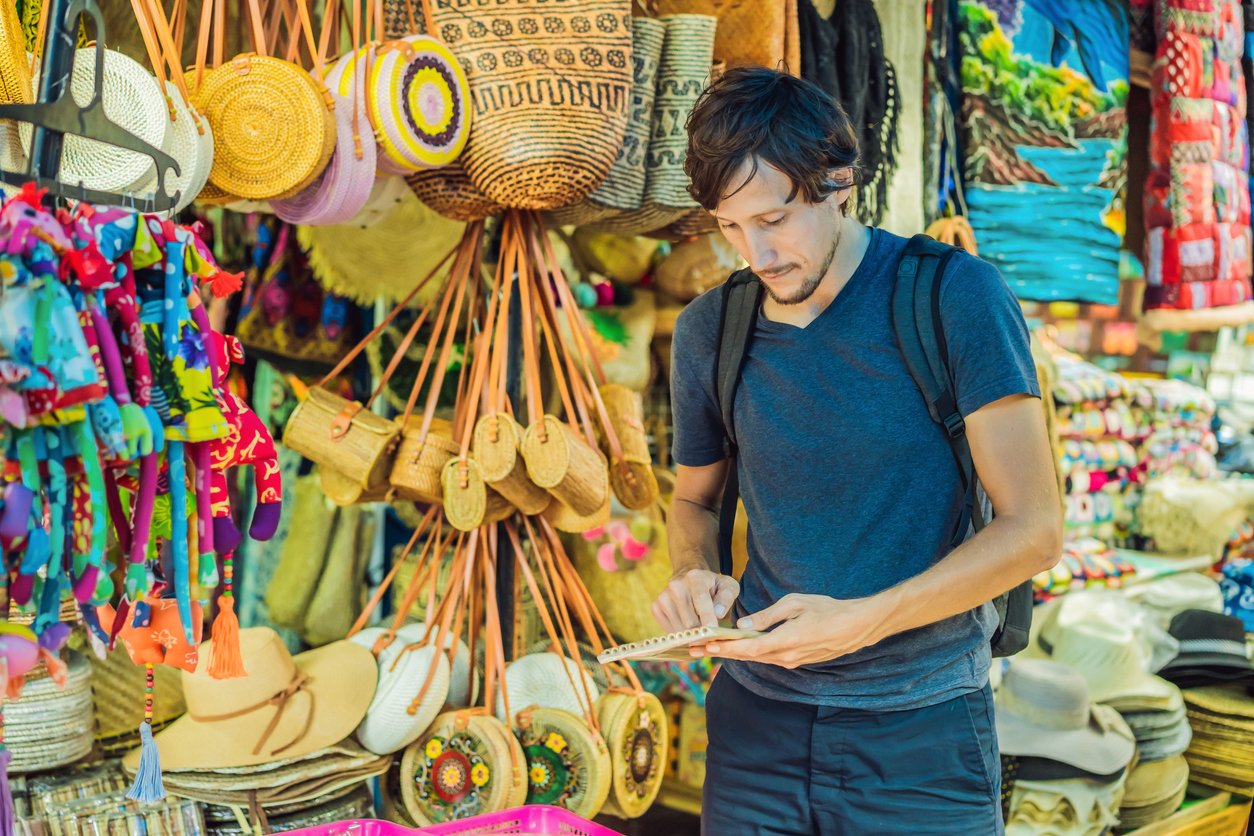 Man with backpack using tablet in colorful outdoor market with hats and bags displayed.