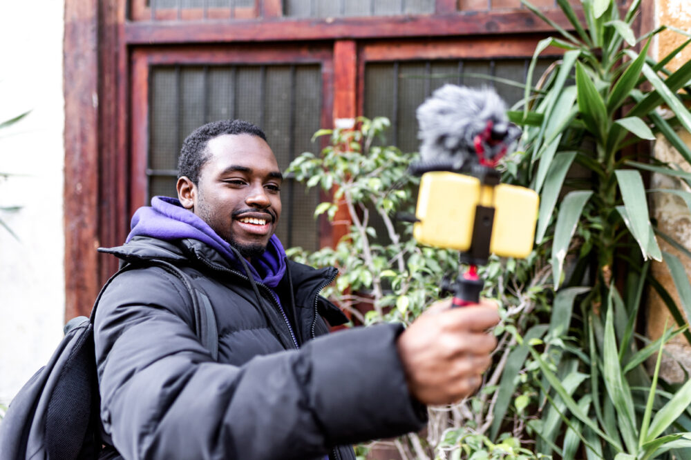 Man in black jacket recording with smartphone and tripod, standing outdoors near plants.