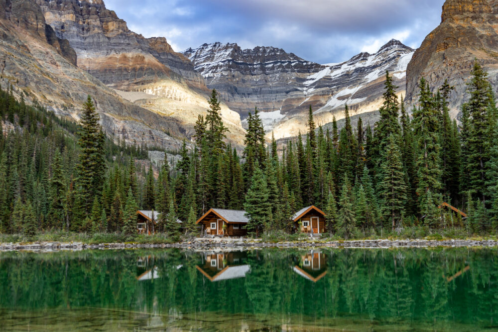 Wooden cabins by a turquoise lake with pine trees and snow-capped mountains in the background.
