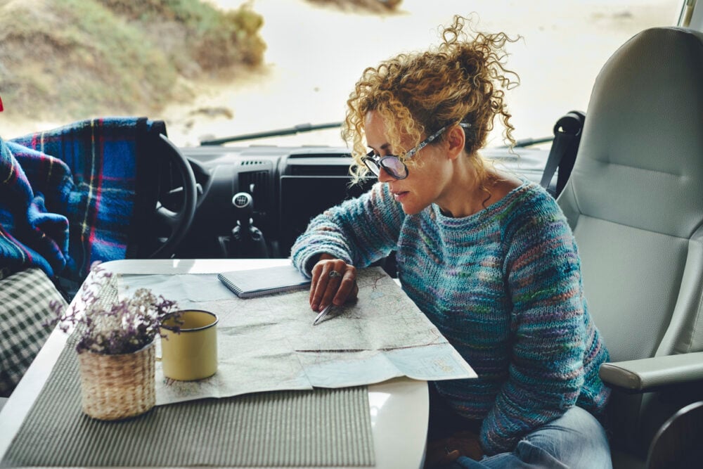 Woman in sunglasses studying map on table inside vehicle, with mug and small basket nearby.