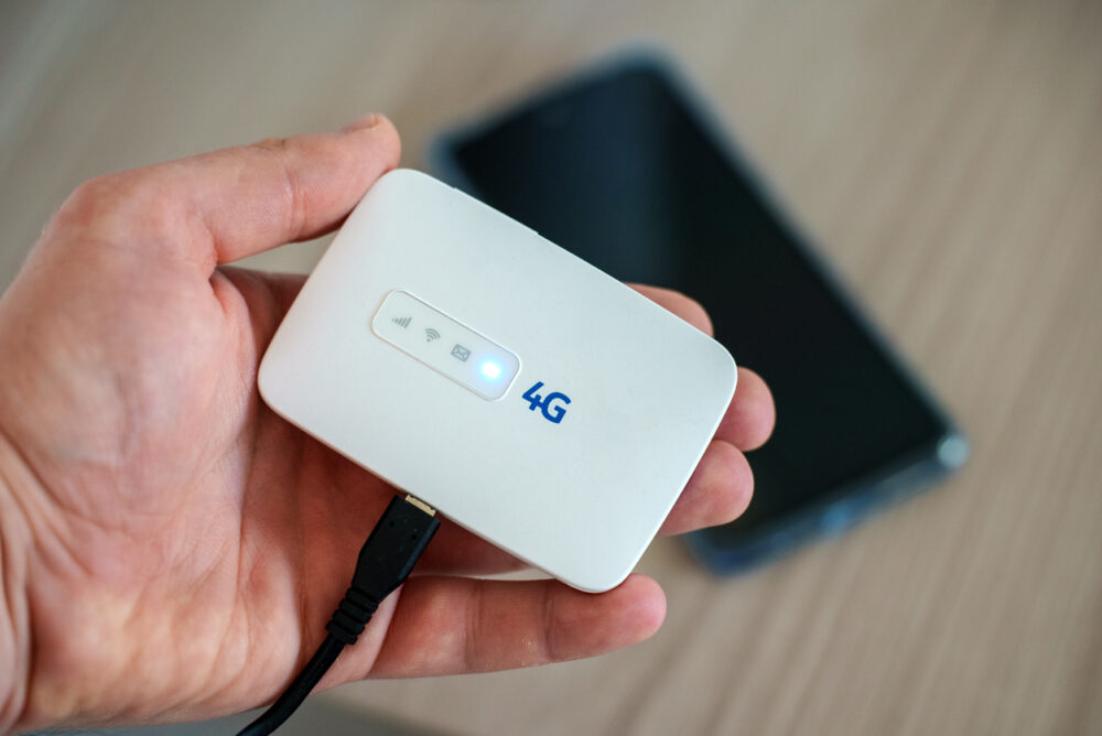 Hand holding a white 4G WiFi router, connected via USB cable, with a smartphone in the background.