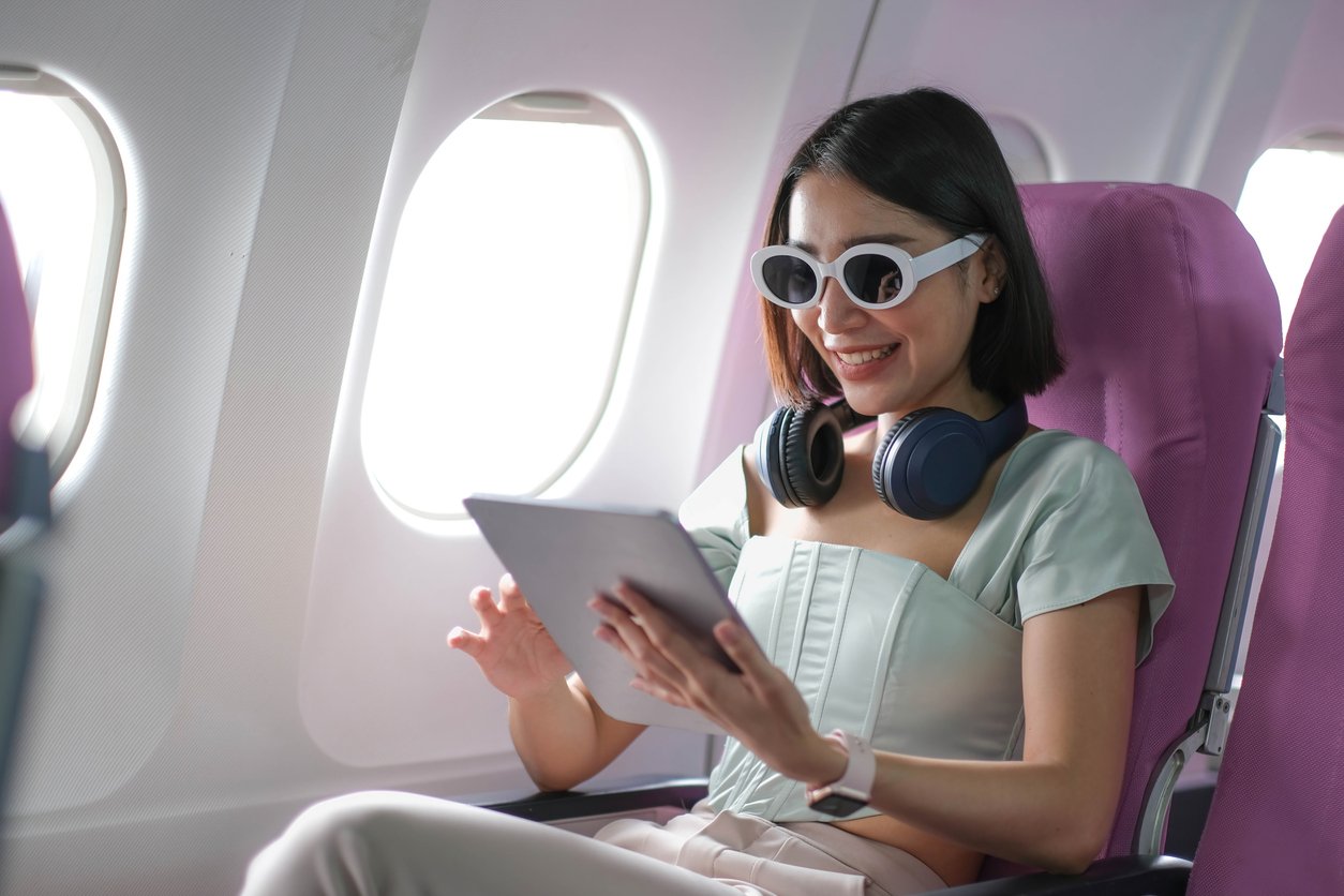 Person on airplane with tablet, wearing headphones and sunglasses, sitting by window in purple seat.