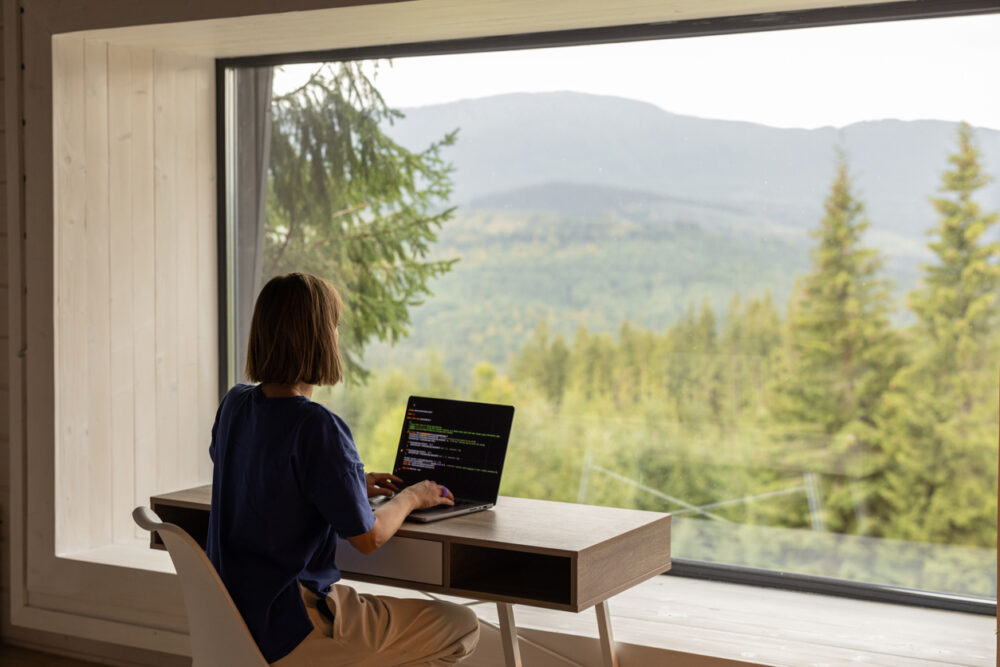 Person typing on a laptop at a desk, overlooking a forested mountain landscape through a large window.