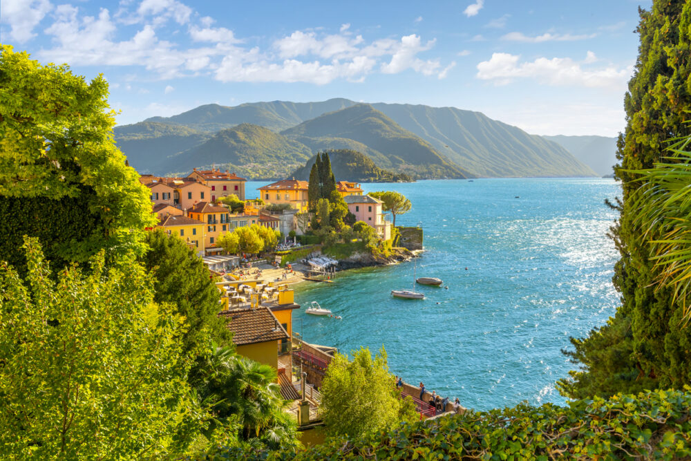 Colorful houses by a bright blue lake, surrounded by green trees and mountains, under a clear sky.