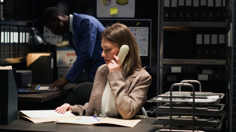 Woman on the phone at a desk, surrounded by folders, with a man working in the background.