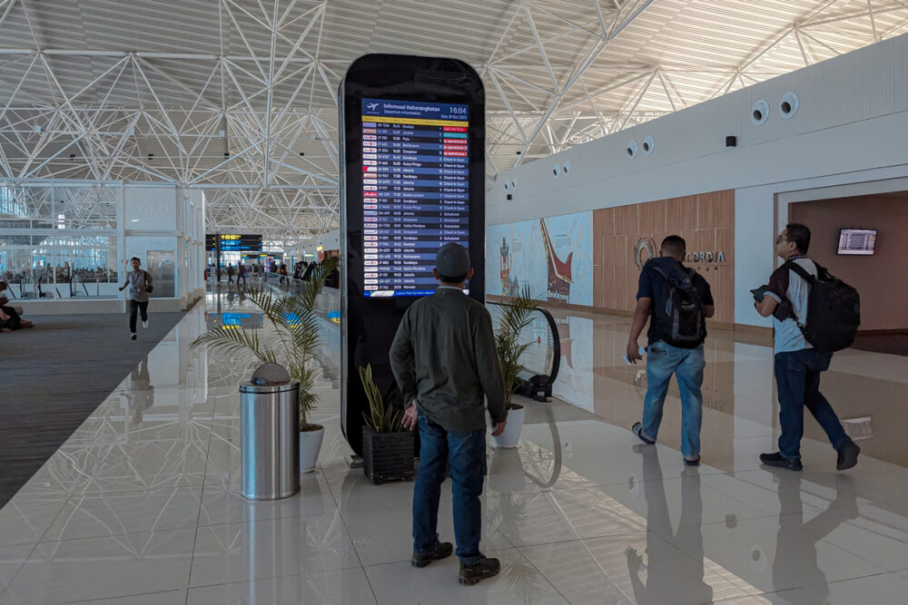 People walking near a digital flight information board in a modern airport terminal with high ceilings.