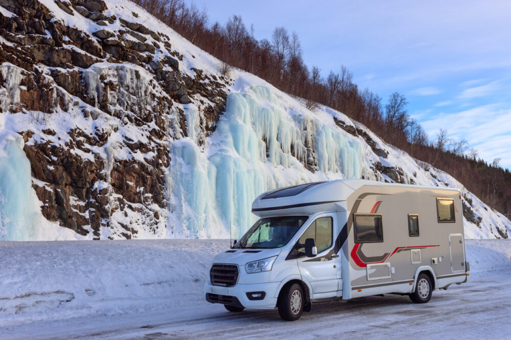 Camper van parked on snowy road beside rocky cliff with frozen waterfalls.