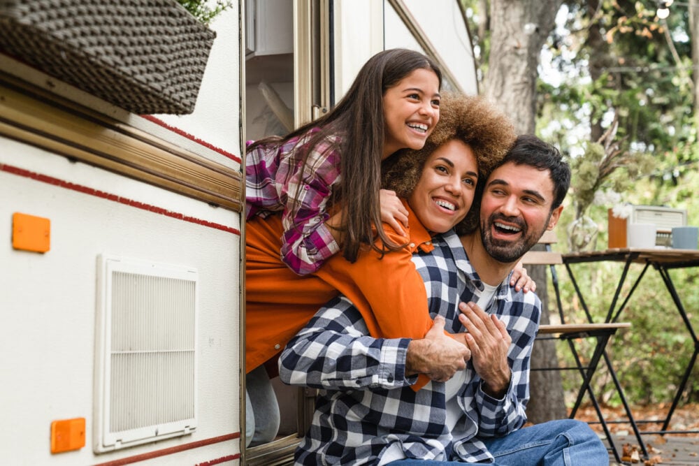 Family of three smiling by a camper, surrounded by trees, with a table and chairs in the background.