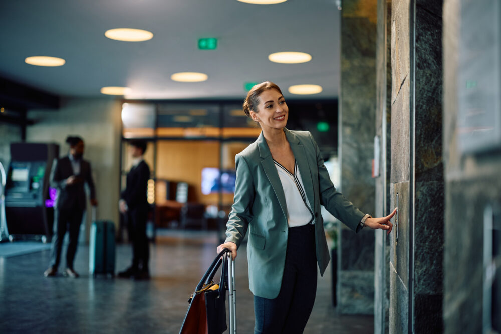 Smiling woman in business attire pressing elevator button in modern lobby with suitcase.