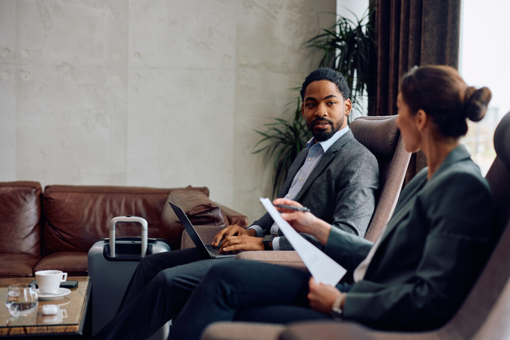 Two business professionals in discussion, one holding papers, seated in a modern office setting with laptop and luggage.