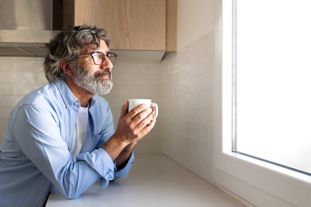 Man with gray beard and glasses holding a mug, looking out of a kitchen window.