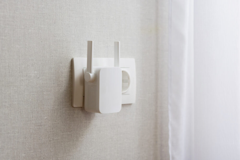 White Wi-Fi extender with antennas plugged into a wall socket on a textured wall background.