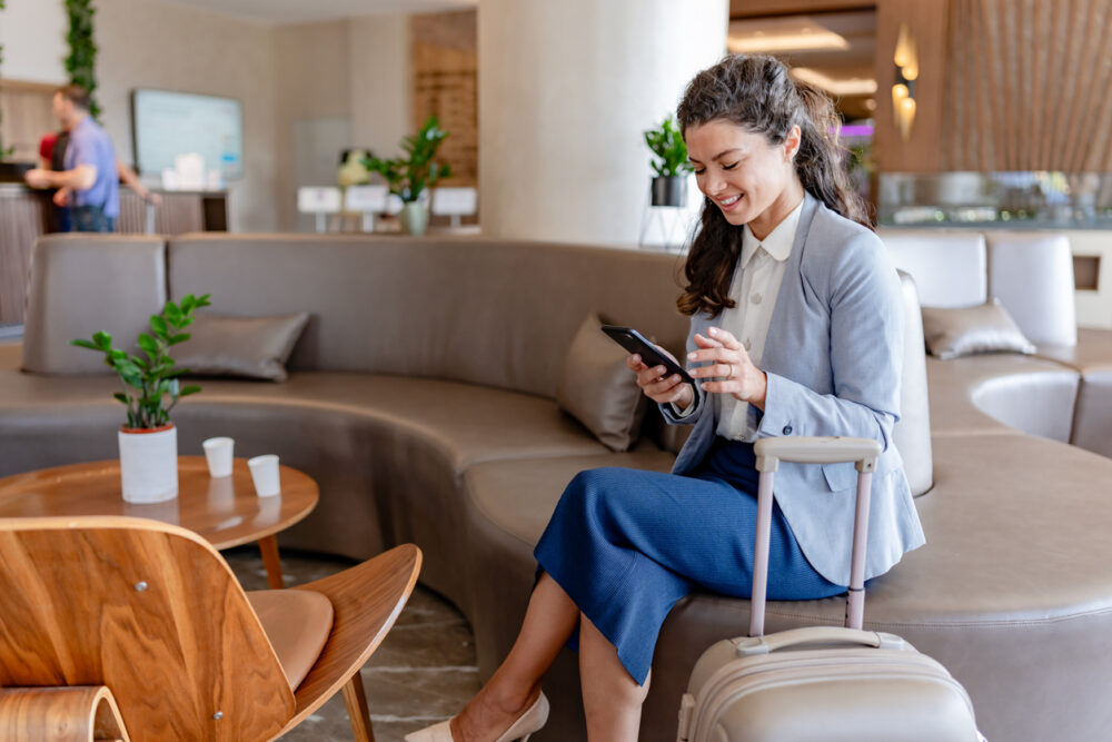 Smiling woman in a blue suit using her phone in a hotel lobby with luggage beside her.