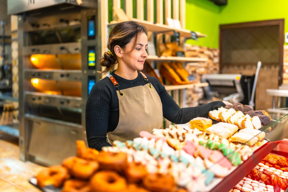 Woman in apron serving assorted pastries in a bakery with ovens in the background.
