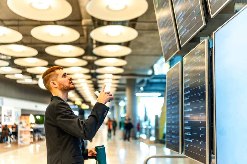 Man in airport pointing at flight information screens, holding a coffee cup, under bright ceiling lights.