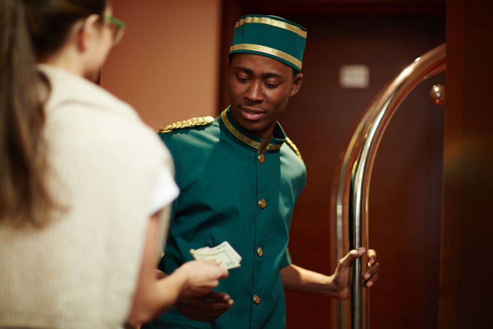 Bellboy in green uniform receiving cash from a woman in a hotel lobby.