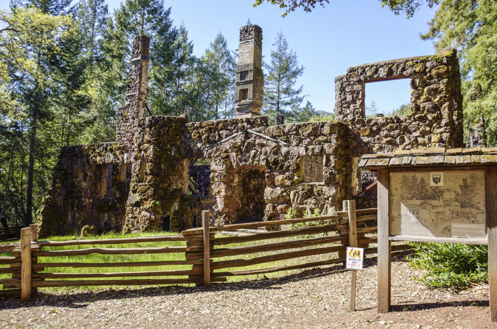 Stone ruins of an old building surrounded by trees and a wooden fence with a small information board.
