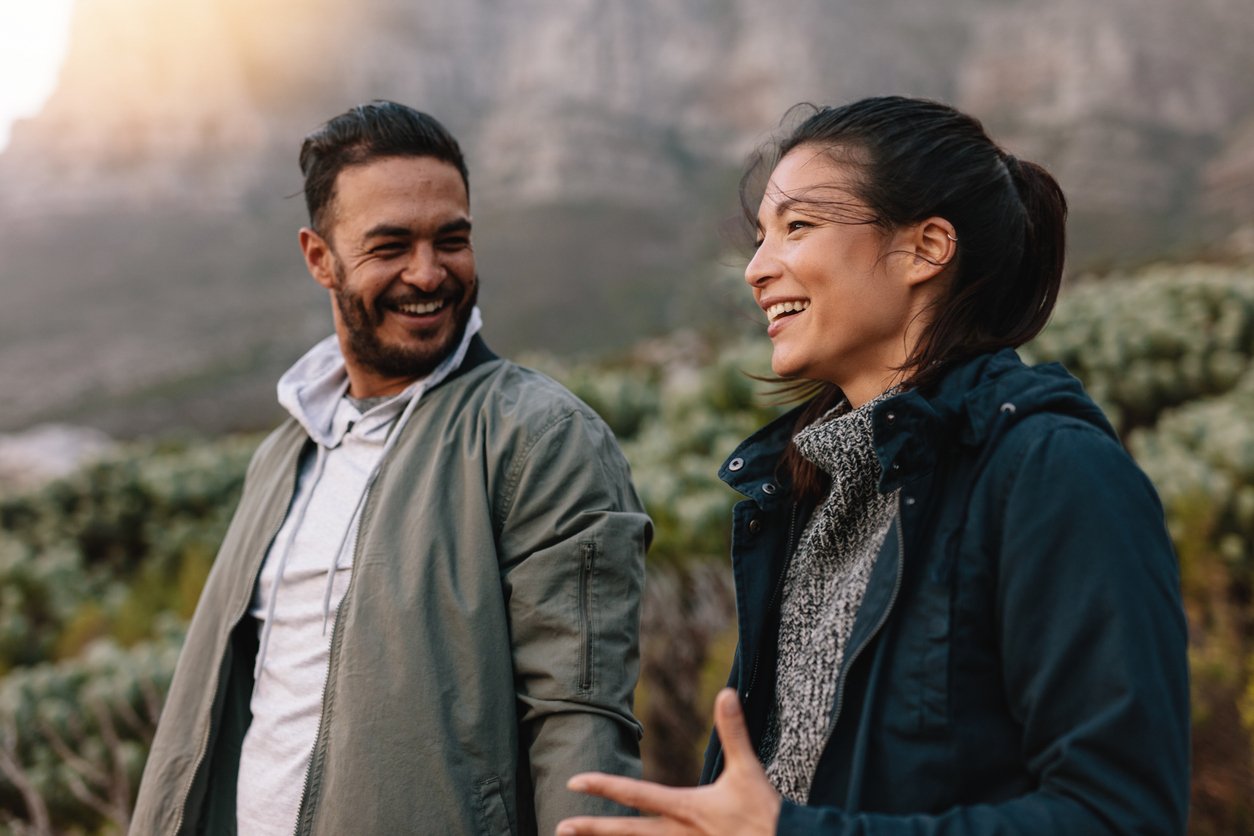 Two people smiling at each other outdoors, with greenery and mountains in the background.