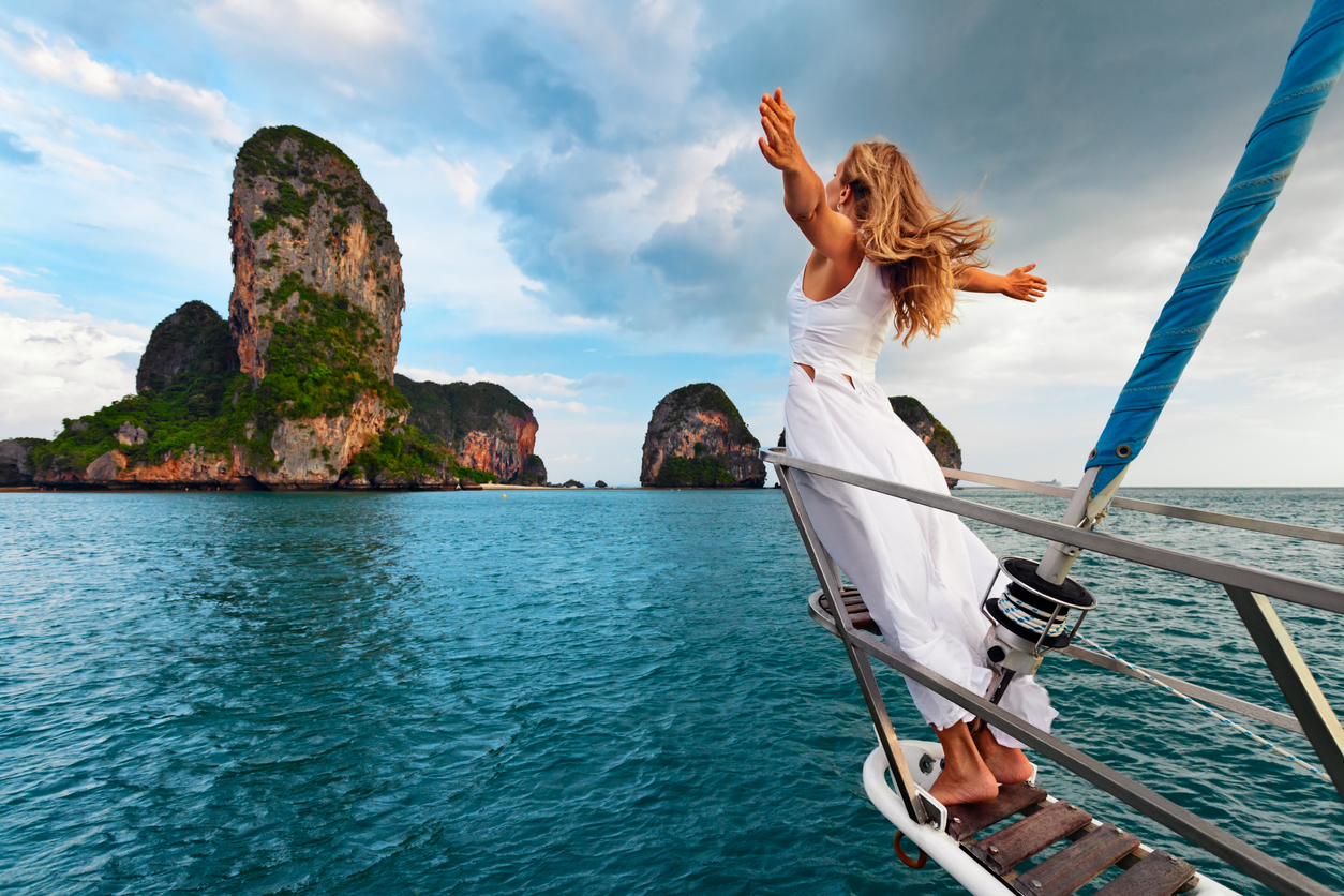 Woman in white dress standing on boat's edge, arms outstretched, with rocky island in background.