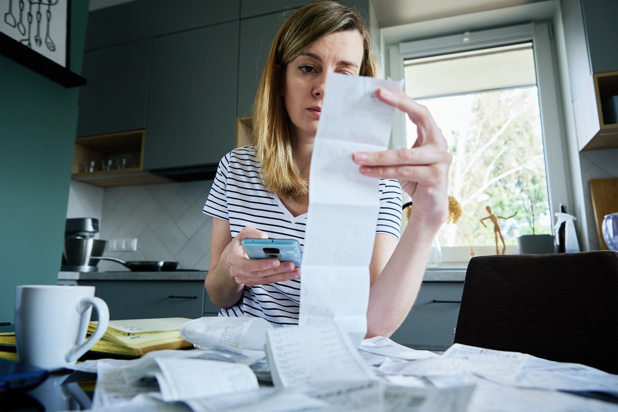 Woman in striped shirt holding smartphone examines long receipt, surrounded by papers on table.