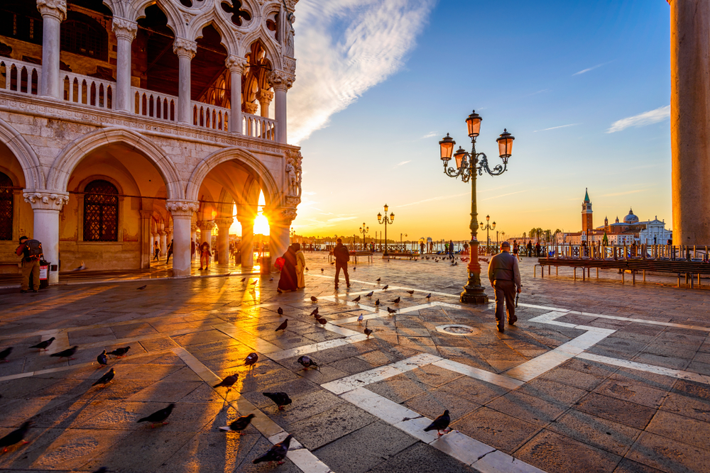 Venice square at sunset, with pigeons, people walking, ornate building, and lampposts casting shadows.