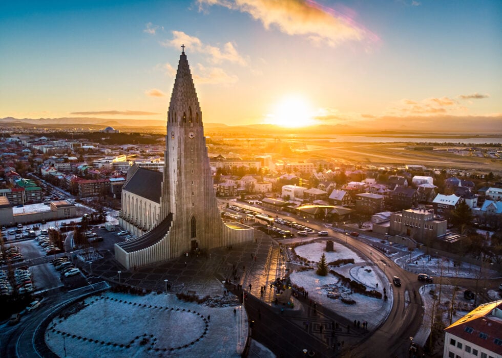 Aerial view of a large church with a pointed tower, surrounded by city buildings at sunset.