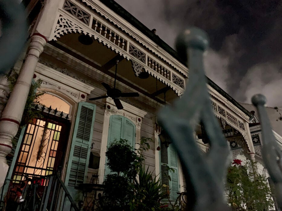 Ornate house at night with shutters, fence, and plants in foreground. Dark cloudy sky above.