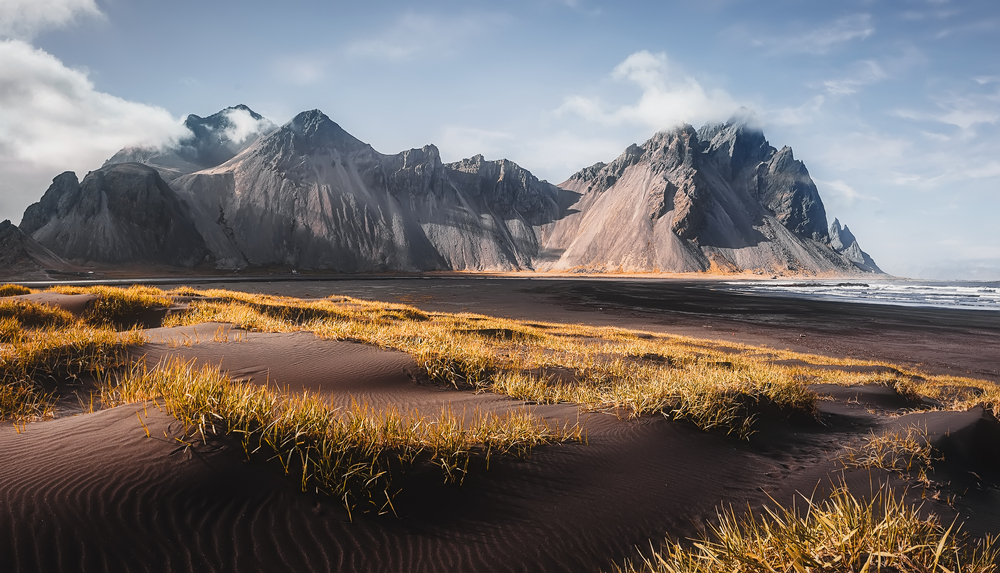 Sandy beach with yellow grass and sharp, rocky mountains under a cloudy sky.