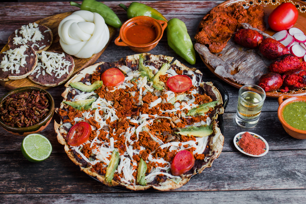 Mexican food spread with tlayuda, chorizo, cheese, chili, lime, tortilla, salsa, and drinks on a wooden table.