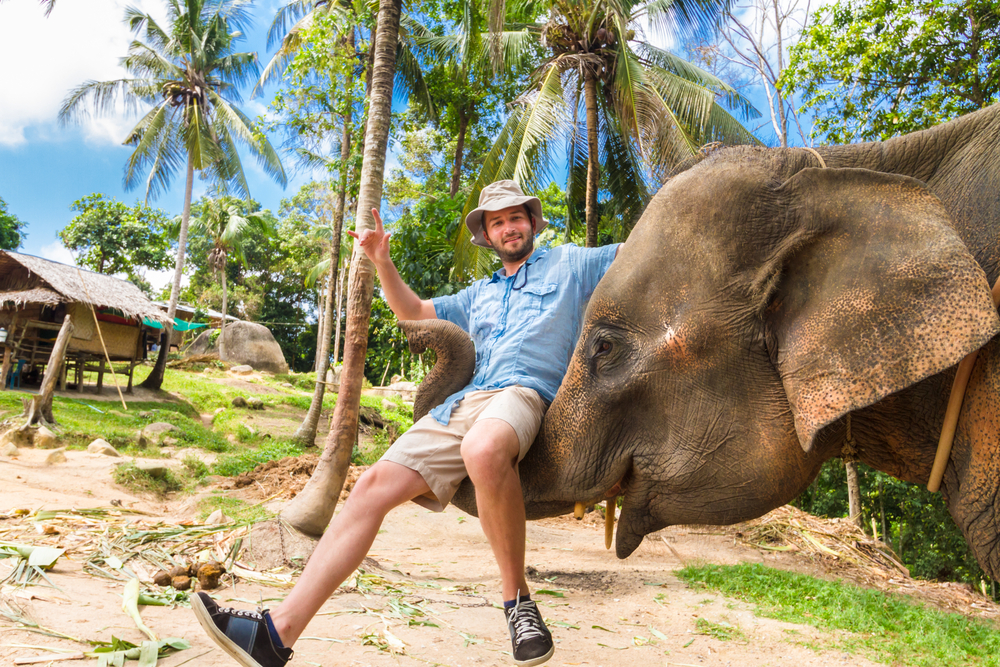Man sitting on an elephant's trunk, smiling, wearing a hat and shorts, with tropical trees in background.