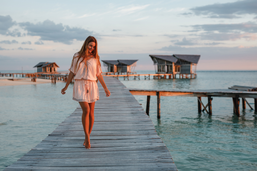 Woman in white dress walking on wooden pier over water at sunset, with huts in the background.