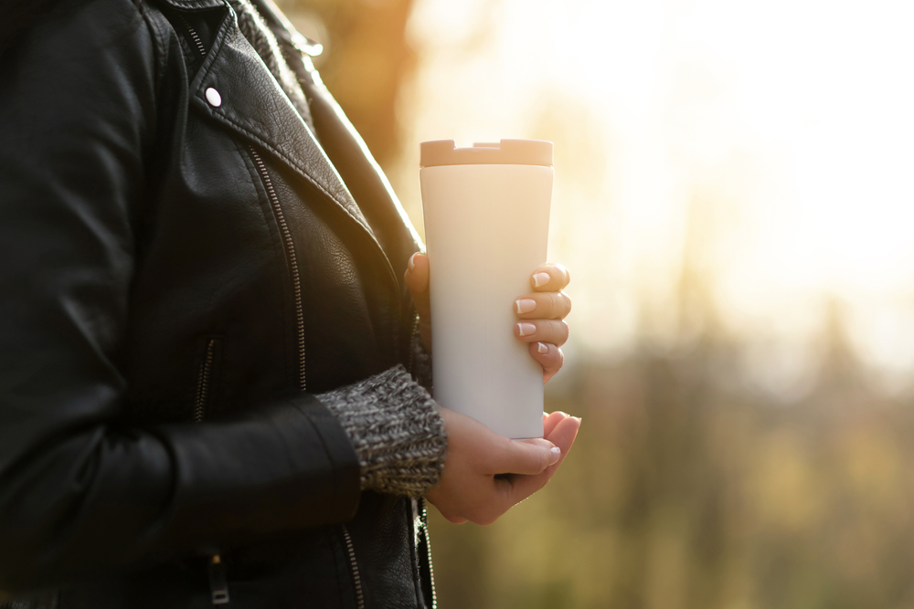 Person holding a white travel mug while wearing a black leather jacket, outdoors.