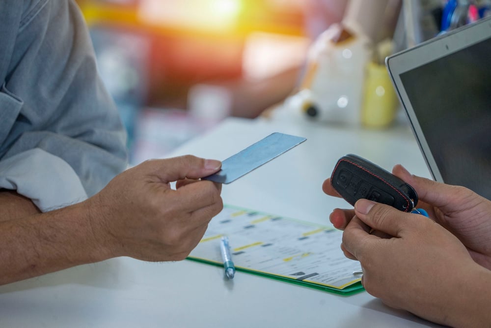 Two people exchange a card and a remote control over a table with a laptop and documents.