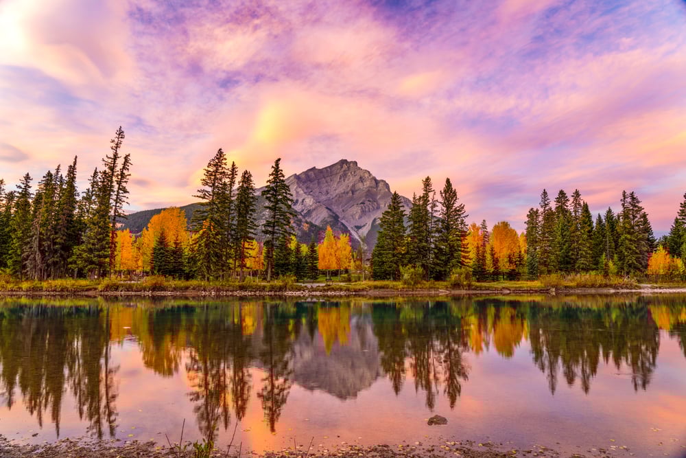A mountain and pine trees reflected in a calm lake under a colorful sunset sky.