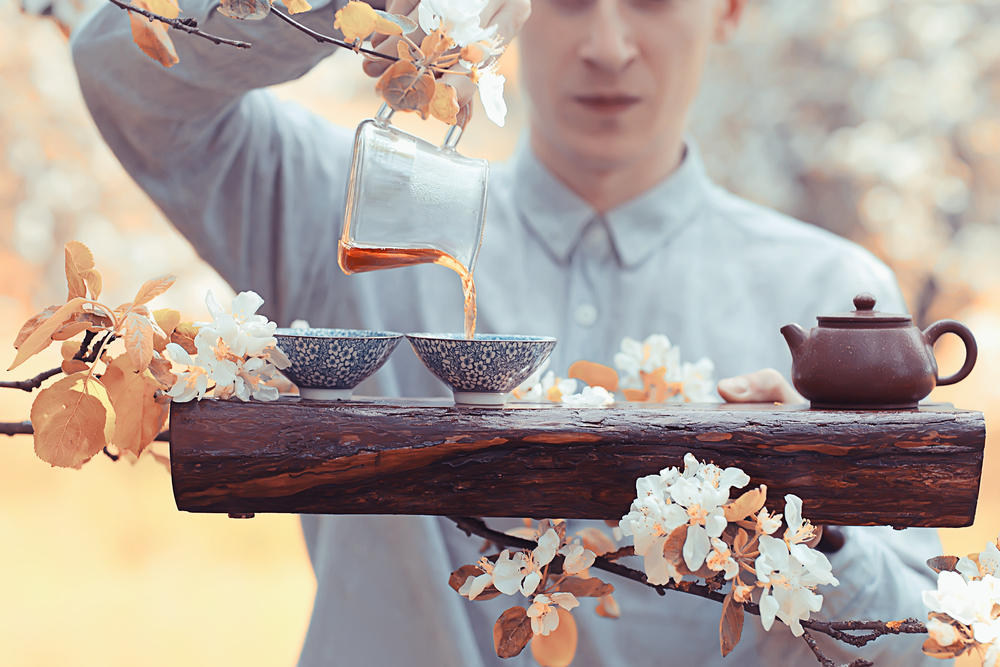 Person pouring tea from glass pitcher into blue cups on a wooden plank among flowering branches.