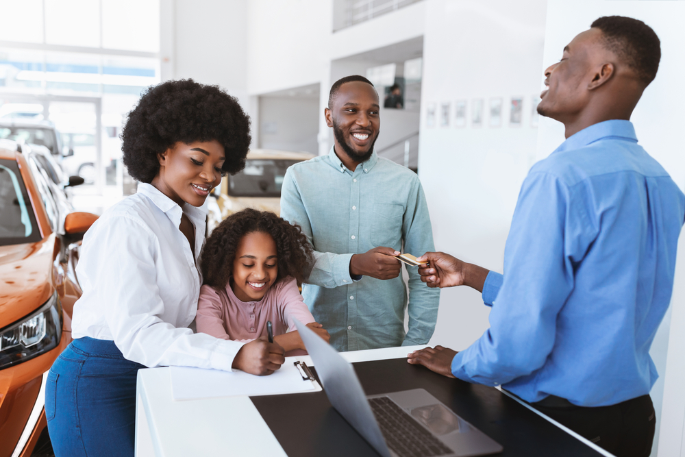 Family signing papers at car dealership, child sitting, adult handing over keys, surrounded by cars.