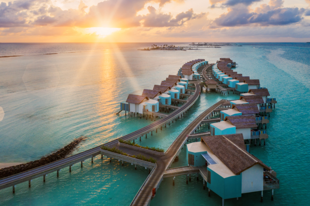 Overwater villas on stilts with wooden roofs, connected by walkways, during sunset over clear ocean water.