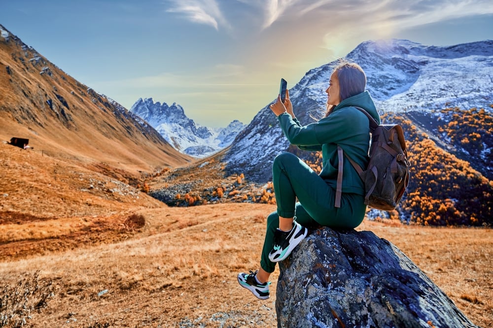 Person in green tracksuit sitting on rock, taking photo with smartphone in mountainous landscape.