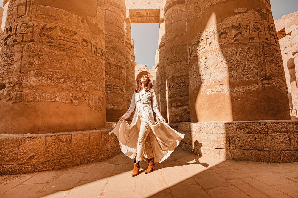 Woman in white dress and hat poses among ancient stone columns with hieroglyphs under clear sky.