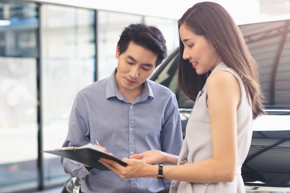 A man and woman review a document on a clipboard inside a car dealership.