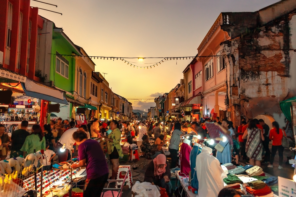Bustling night market with colorful buildings, street vendors, and shoppers under decorative string lights at sunset.