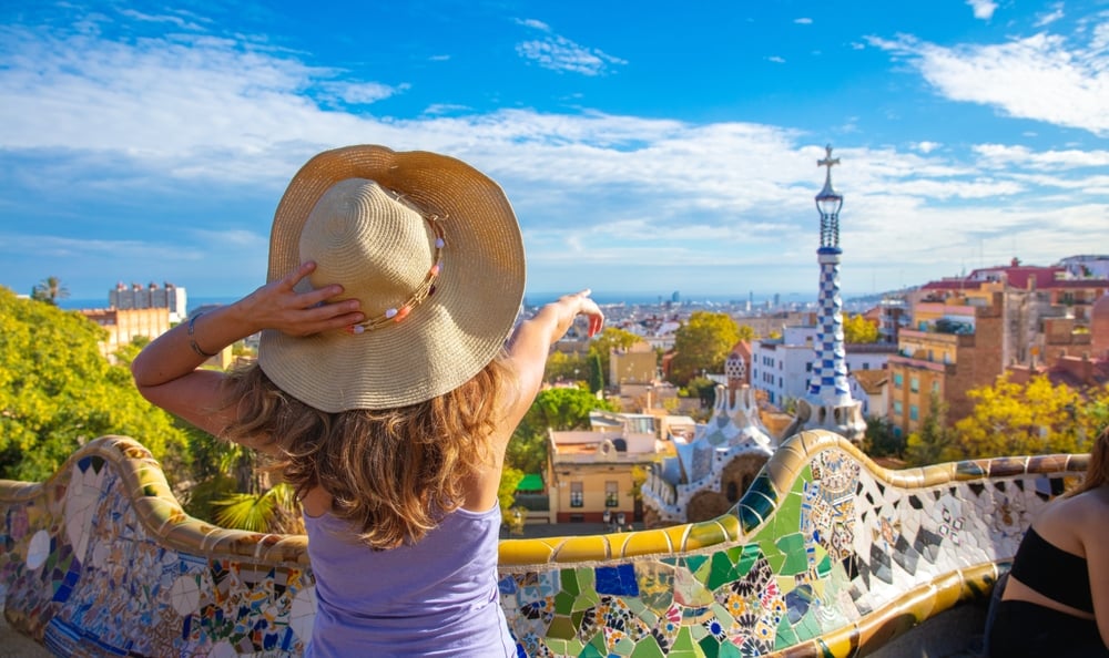 A woman in a hat points towards colorful buildings and a mosaic bench at Park Güell, Barcelona.