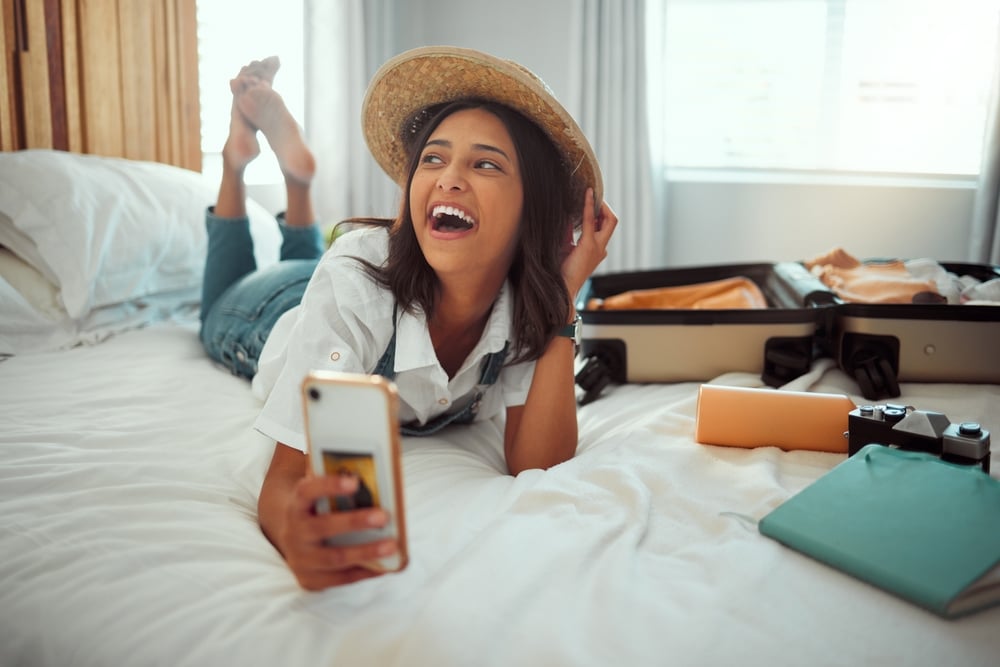 Woman in a straw hat lying on a bed, holding a smartphone, with an open suitcase nearby.