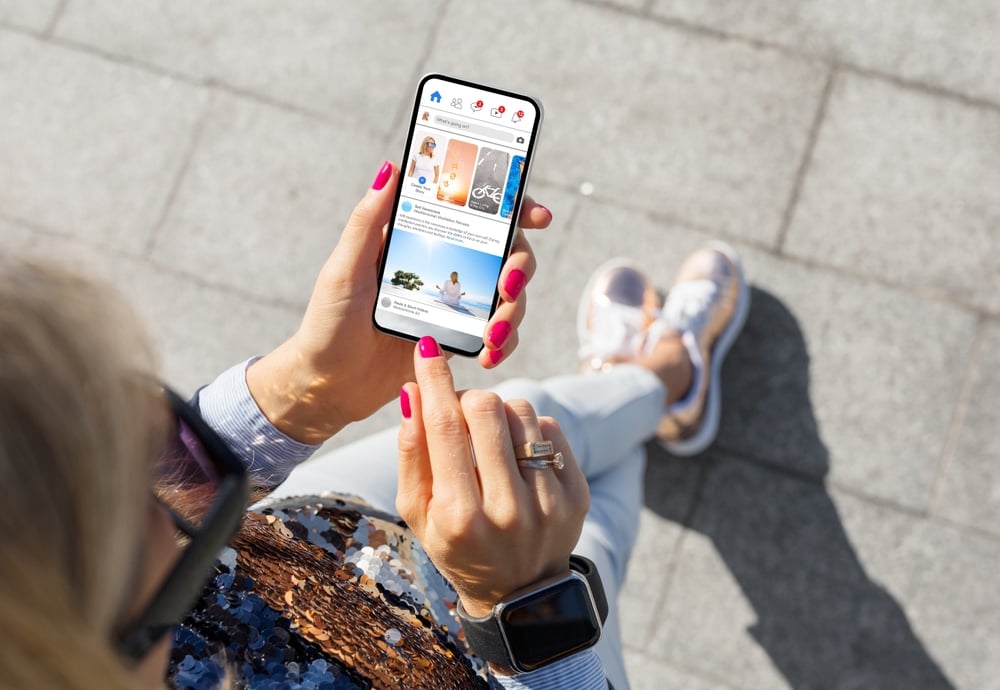 A person with pink nails taps a smartphone, displaying a social media app, while sitting on pavement.