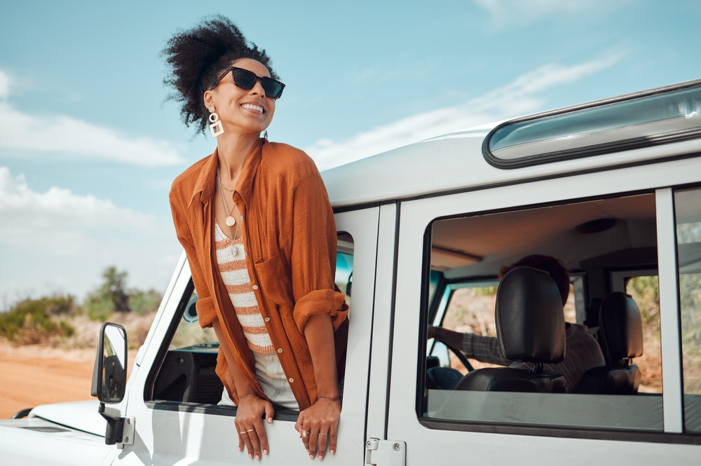 Woman wearing sunglasses, leaning out of car window, smiling under clear blue sky.
